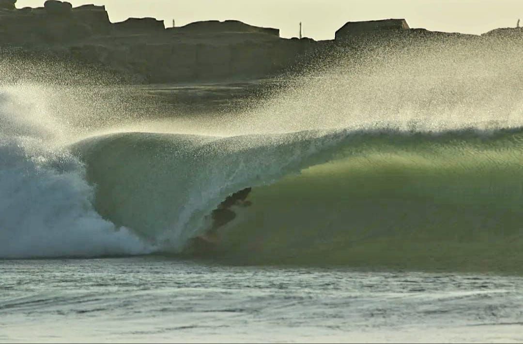 A dynamic wave curls gracefully in the ocean, displaying a spectrum of green and blue hues. The mist from the water catches the light, creating a soft glow. Rugged rock formations are visible in the background, framing the scene.