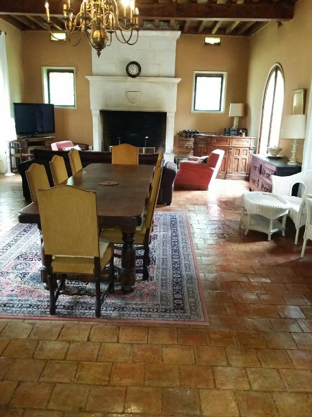 A spacious dining area features a long wooden table surrounded by eight chairs, with a patterned area rug beneath. A stone fireplace is centrally located, with a clock mounted above. Nearby, a cozy seating area includes an armchair and a two-seater couch, illuminated by natural light.
