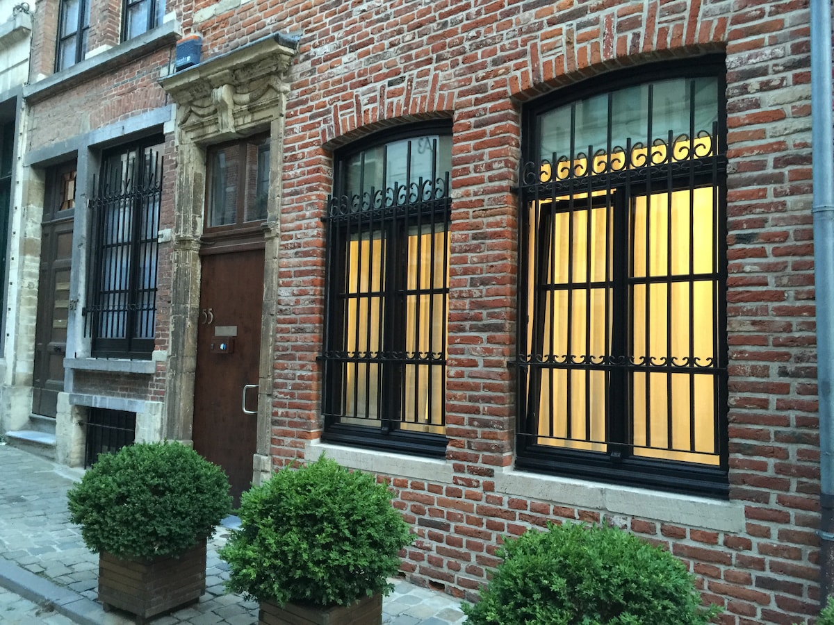 A charming brick facade is featured, showcasing two large windows with decorative black iron grilles. Soft, warm light glows from within, illuminating the space. Flanking the entrance, small topiary plants in wooden planters add greenery to the setting.