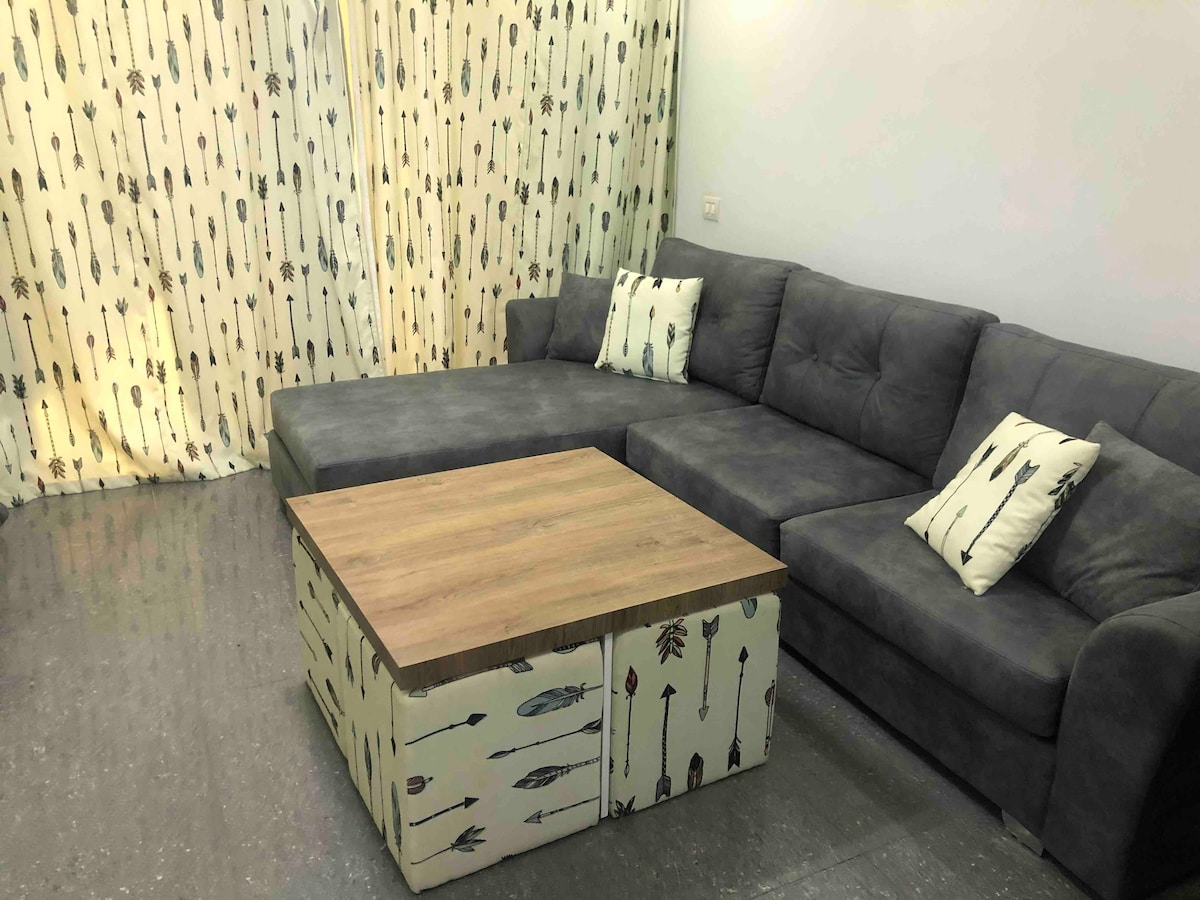 A spacious seating area features a plush grey sectional sofa with decorative pillows. A coffee table, designed with an arrow motif, occupies the center, complemented by softly patterned curtains that provide a touch of elegance to the room.