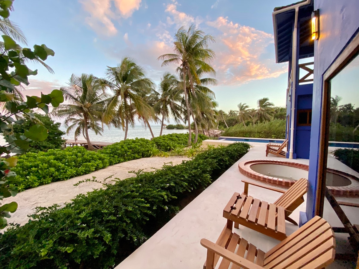 A peaceful outdoor space features wooden lounge chairs positioned near lush greenery and a view of the beach. The scene is framed by swaying palm trees under a soft sunset sky, providing a serene ambiance for relaxation.