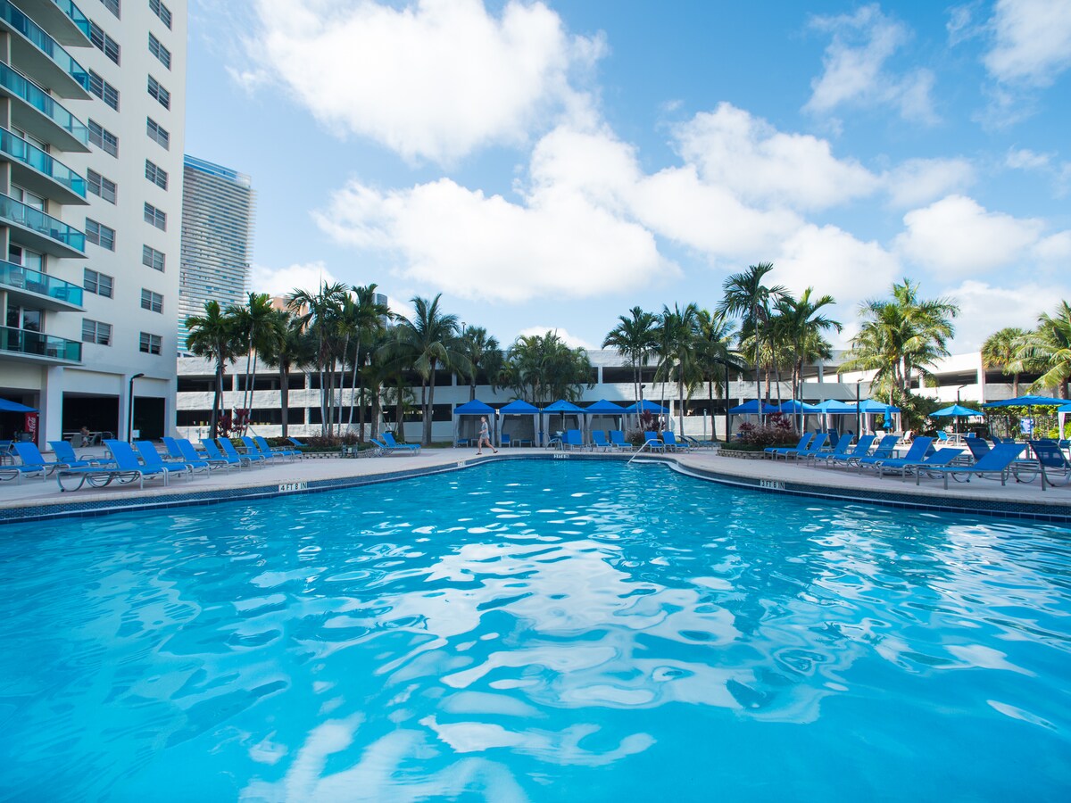 A large outdoor heated swimming pool is surrounded by lounge chairs and palm trees. Bright blue skies enhance the refreshing atmosphere, while shaded gazebos are positioned along the pool’s edge, offering relaxation options for guests.