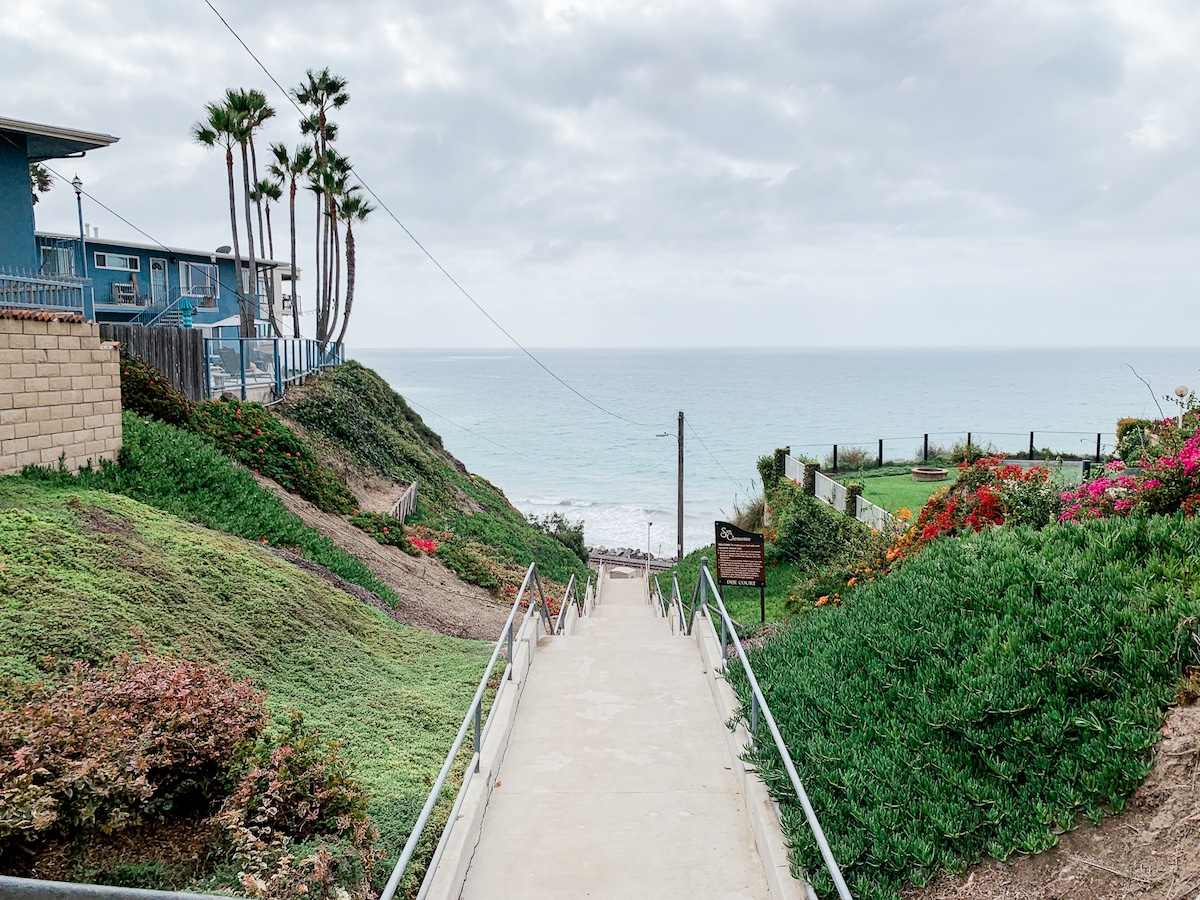 A pathway leading down towards the ocean is framed by lush greenery and vibrant flowers. Palm trees line the side, while a cloudy sky looms overhead. The calming sound of waves can be imagined just beyond the horizon.