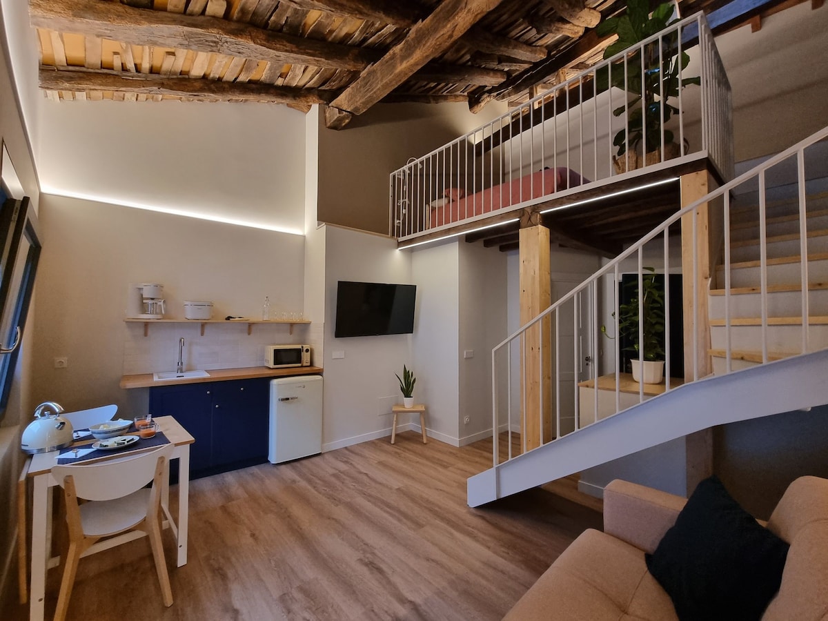 A spacious open-plan living area features light wood accents, a kitchenette with a microwave, and a cozy sofa. A staircase leads to an upper sleeping area, where a double bed is visible. Natural light illuminates the space, creating a calm atmosphere.