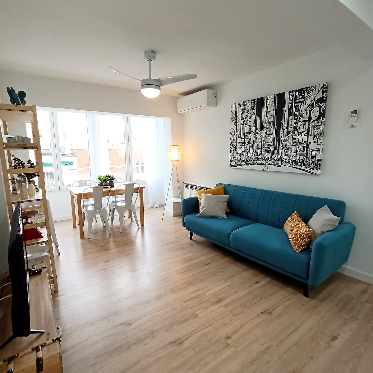 The living area showcases a modern design with a bright blue sofa and a light-colored dining table set for four. Large windows fill the space with natural light, complemented by a ceiling fan and contemporary artwork on the wall.