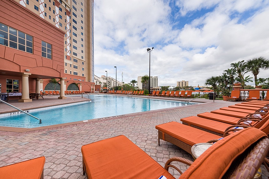 An outdoor pool area is featured, surrounded by lounge chairs with orange cushions. The pool reflects the sky, and palm trees provide a tropical ambiance. The building in the background adds a modern touch, while the pool area invites relaxation in an inviting setting.