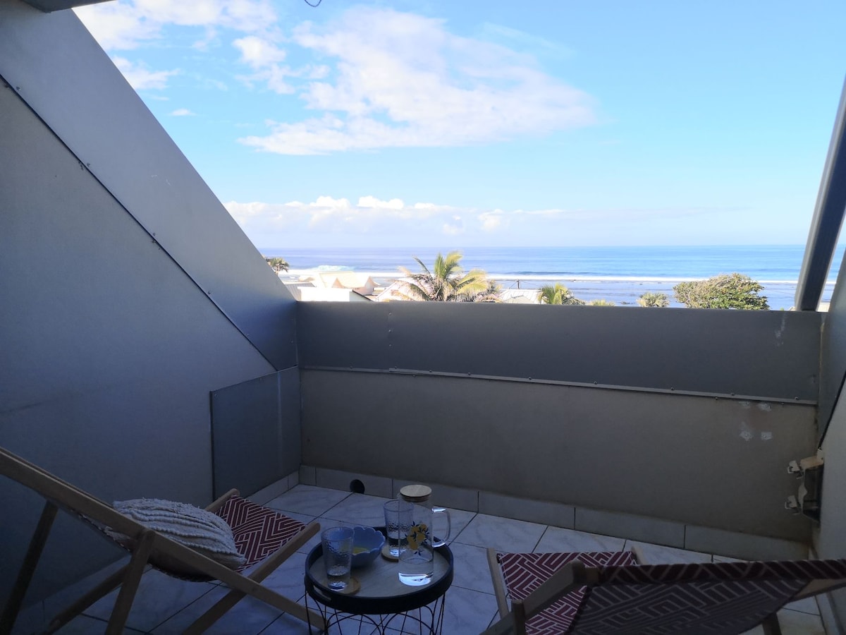 A balcony is observed with a stunning view of the sea and sky, featuring two lounge chairs. A small table displays glassware, creating an inviting space for relaxation. The surrounding area includes glimpses of palm trees and the coastline in the distance.