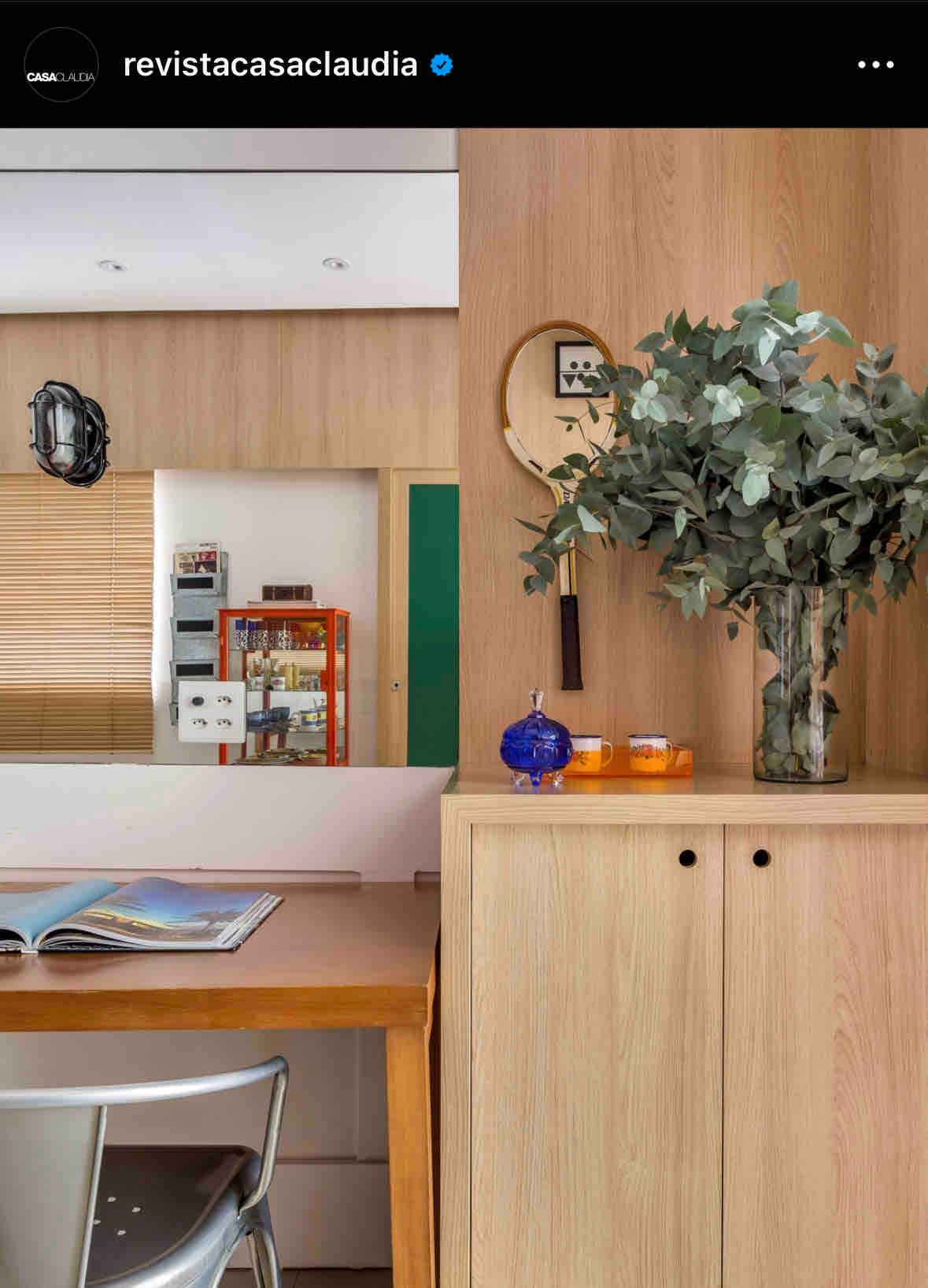 A wooden console features a decorative vase filled with greenery. An open magazine lies on a small desk, alongside a blue glass decorative piece and a few candles. A mirror reflects a glimpse of the kitchen area, contributing to a spacious feel.
