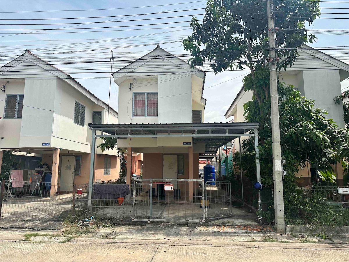 The image showcases a single-story row house in a residential setting. A covered parking area is visible in the foreground, with plants lining the entrance. Neighboring houses can be seen in the background, adding to the community feel.