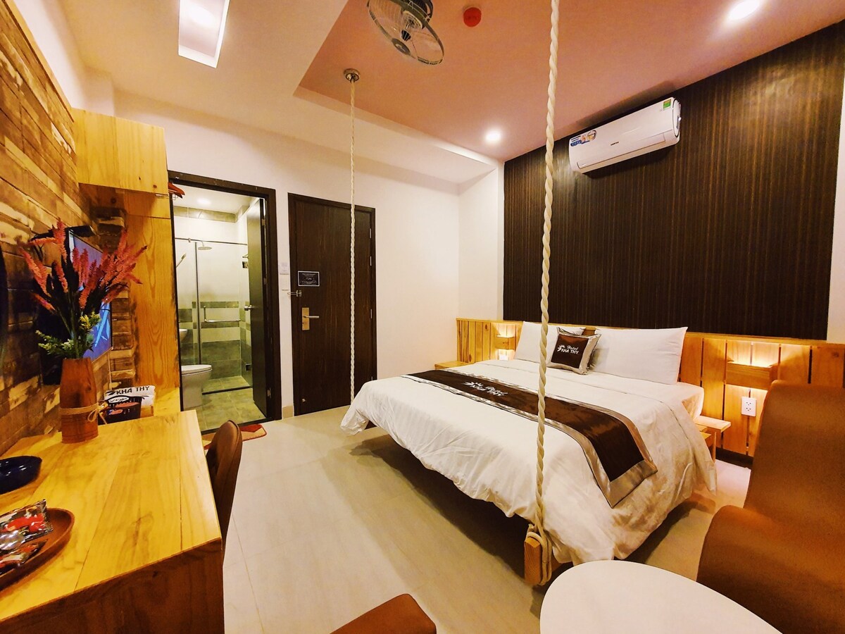 A modern room features a spacious bed with a dark accent wall behind it. Natural wooden elements are highlighted in the furnishings, including a desk and seating area. A bathroom entrance is visible in the background, with a colorful arrangement of flowers positioned on the desk.