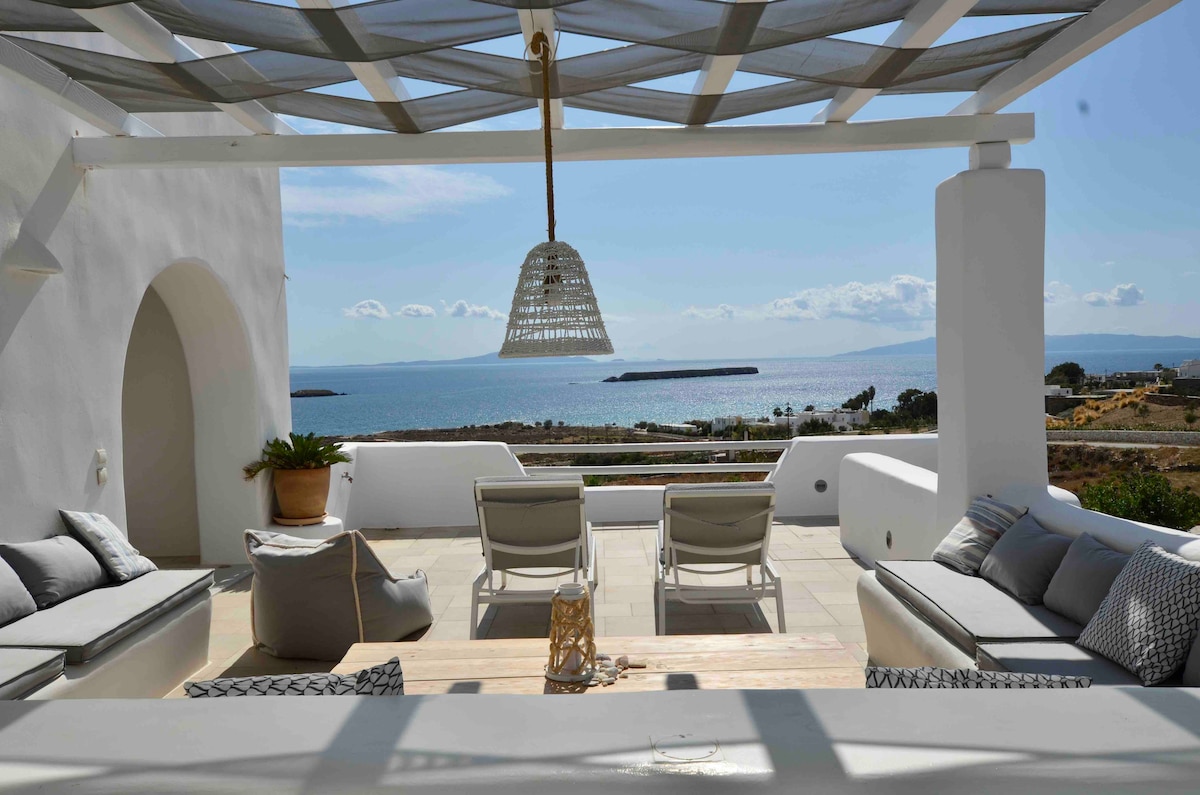 A spacious outdoor lounge area features comfortable seating arranged along the edges, with light cushions against a backdrop of panoramic sea views. A wooden table stands at the center, while a pendant light hangs above, adding to the inviting atmosphere.