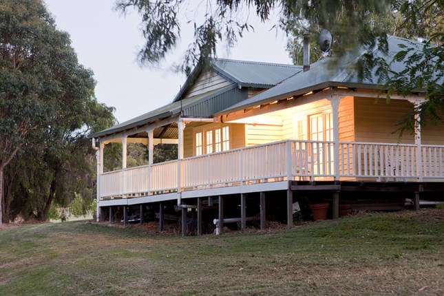 The cottage features a spacious veranda that wraps around the exterior, inviting guests to enjoy the serene surroundings. Soft lighting illuminates the entrance, highlighting the wooden paneling and light-colored trim of the facade. The structure is set atop sturdy supports, surrounded by lush greenery.