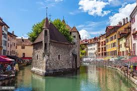 A historic building with a pointed roof stands on a small island in the water, surrounded by quaint, colorful buildings. Lush greenery is visible atop the structure, with boats and outdoor seating along the water's edge. Bright blue skies complement the scene.