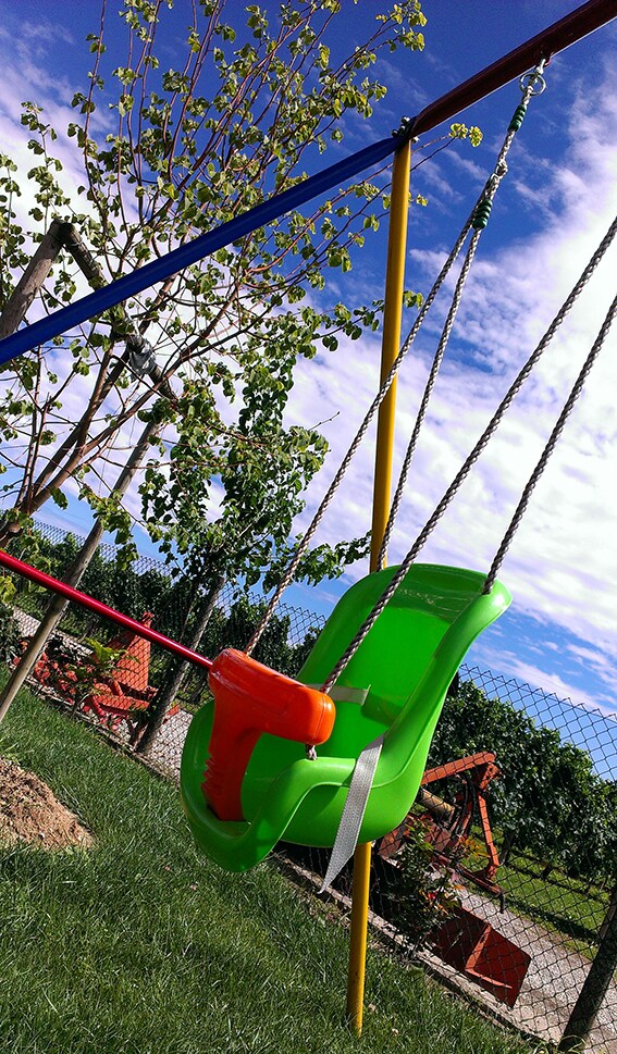 A bright green baby swing is positioned within a grassy area, secured with safety straps. Behind the swing, trees provide a natural backdrop against a blue sky with scattered clouds. The surroundings are designed for family-friendly outdoor enjoyment.