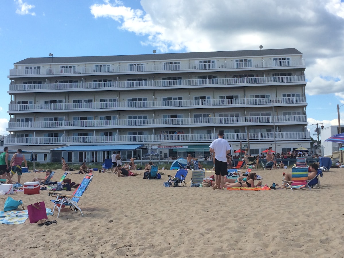 A four-story oceanfront condo building features multiple balconies that overlook a sandy beach. Sunbathers and beach chairs are spread across the shoreline, with a vibrant atmosphere enhanced by people enjoying the coastal setting under a partly cloudy sky.
