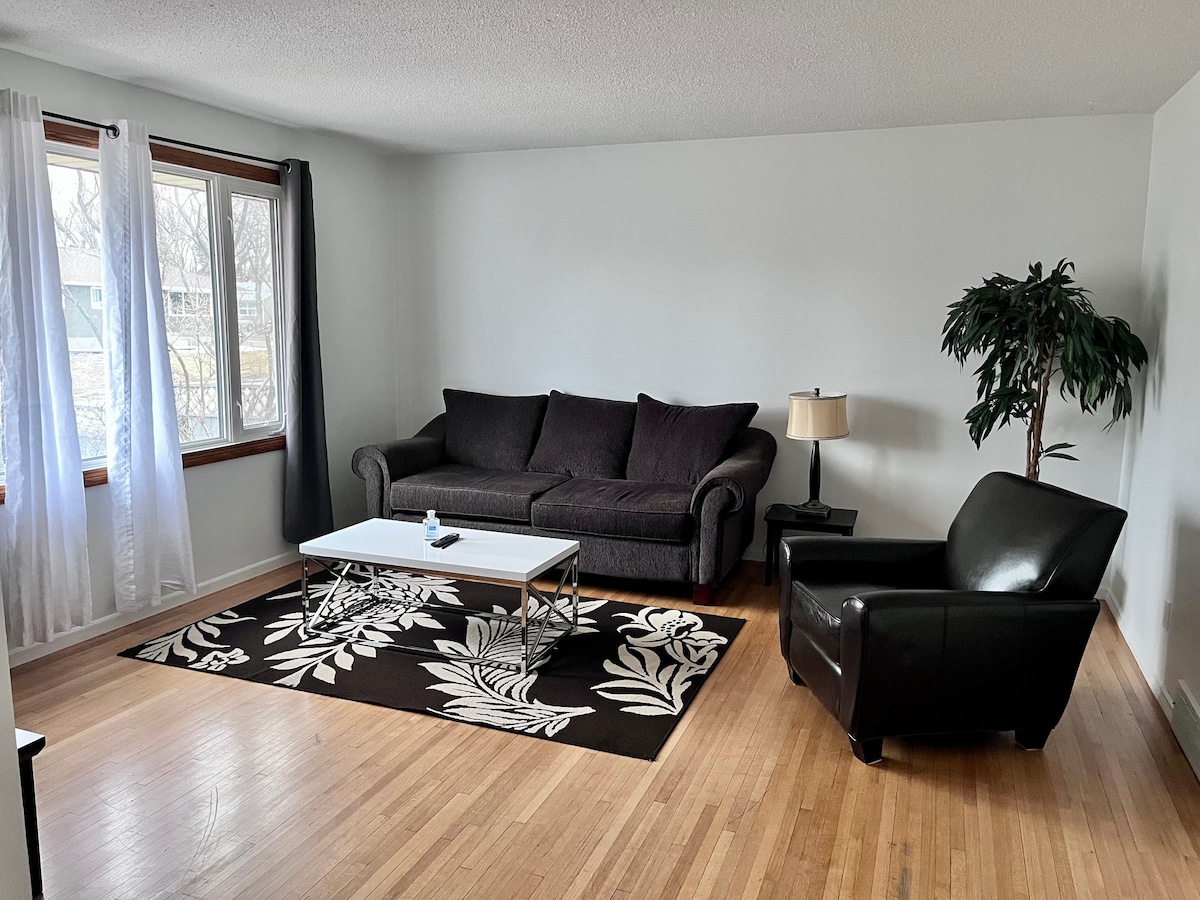 A comfortable living room presents a dark sofa and an armchair beside a small, modern coffee table. Natural light enters through large windows, adorned with sheer white curtains, highlighting the light wood flooring and a leaf-patterned area rug.