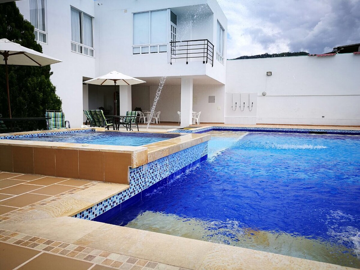 A large outdoor pool area features a spacious swimming pool with varying depths, complemented by a smaller wading pool. Sun loungers and umbrellas are arranged nearby against a backdrop of white walls and large windows, creating an inviting space for relaxation.