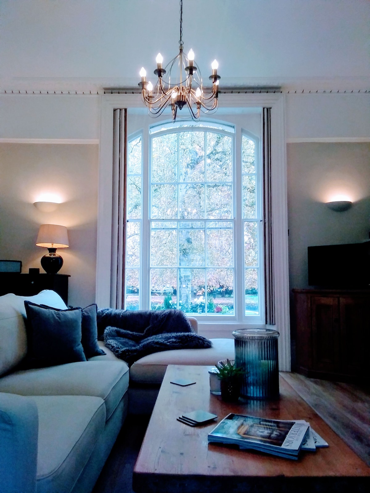 A spacious living area is depicted, featuring a cozy white sofa adorned with dark cushions. A wooden coffee table sits in front, showcasing magazines. Natural light floods the room through a large arched window, offering views of greenery outside, complemented by soft wall lights.