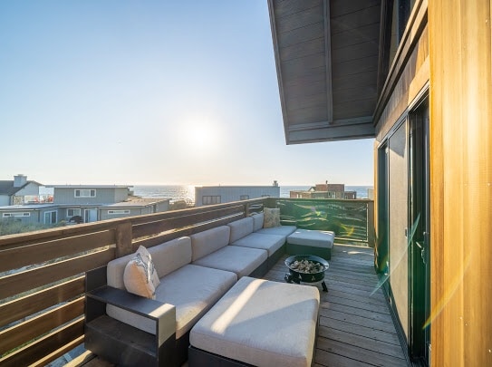 A spacious outdoor seating area features a sectional sofa arranged around a circular fire pit on the deck. The setting offers expansive views of the ocean, with sunlight reflecting off the water and a clear sky above.