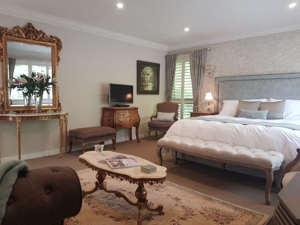 A refined guest suite features a spacious bed dressed in soft linens, flanked by elegant side tables. A vintage-style mirror is positioned above a decorative console table, while two armchairs create a cozy seating area. Light streams through the window, enhancing the warm tone of the decor.