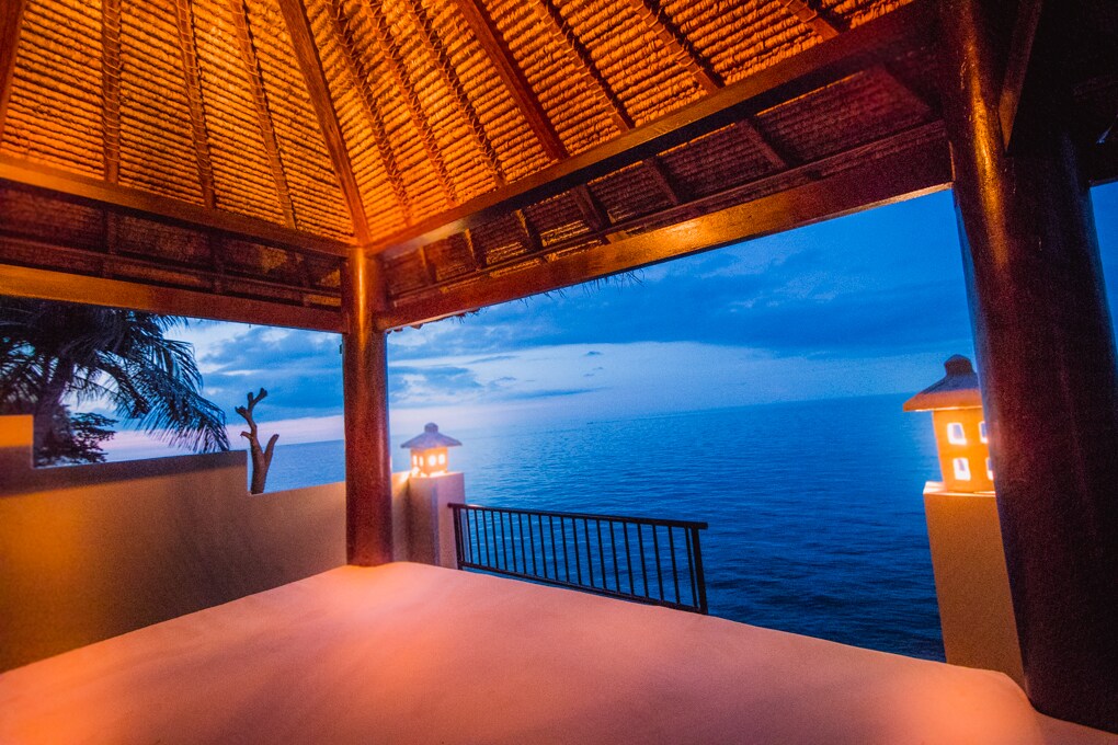 A cozy gazebo is featured overlooking the calm sea at twilight. Soft lighting from lanterns casts a warm glow, enhancing the tranquil ambiance. The thatched roof provides shade, while the gentle waves create a serene backdrop.