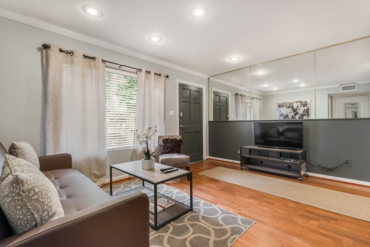 A cozy living area features a brown sofa with decorative pillows, accompanied by a patterned area rug. A dark coffee table is centered in front of the sofa, with a flat-screen TV positioned on a media unit against the wall. Natural light enters through the window, framed by light curtains.