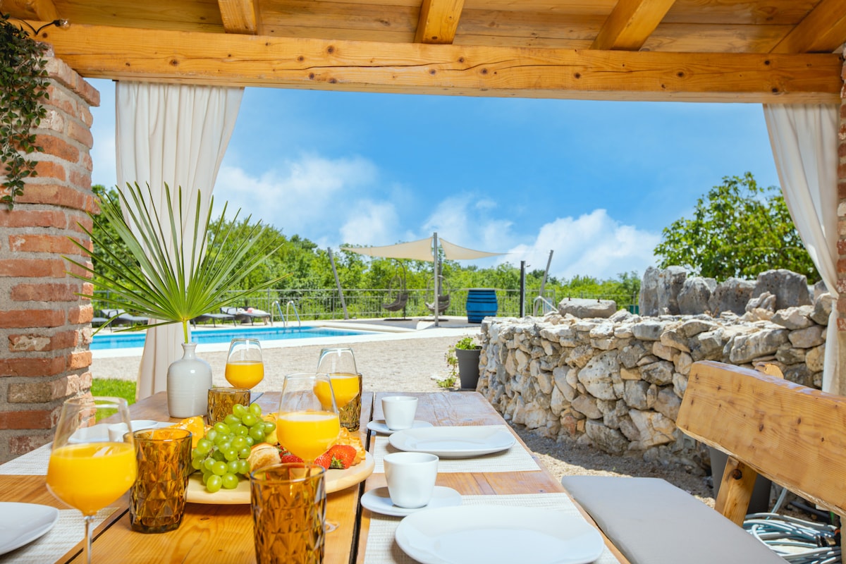 An outdoor dining area is presented with a wooden table set for breakfast, featuring glasses of orange juice and an assortment of fruits. A serene view of the pool and surrounding greenery is framed by the wooden beams and draped fabric of the pavilion.