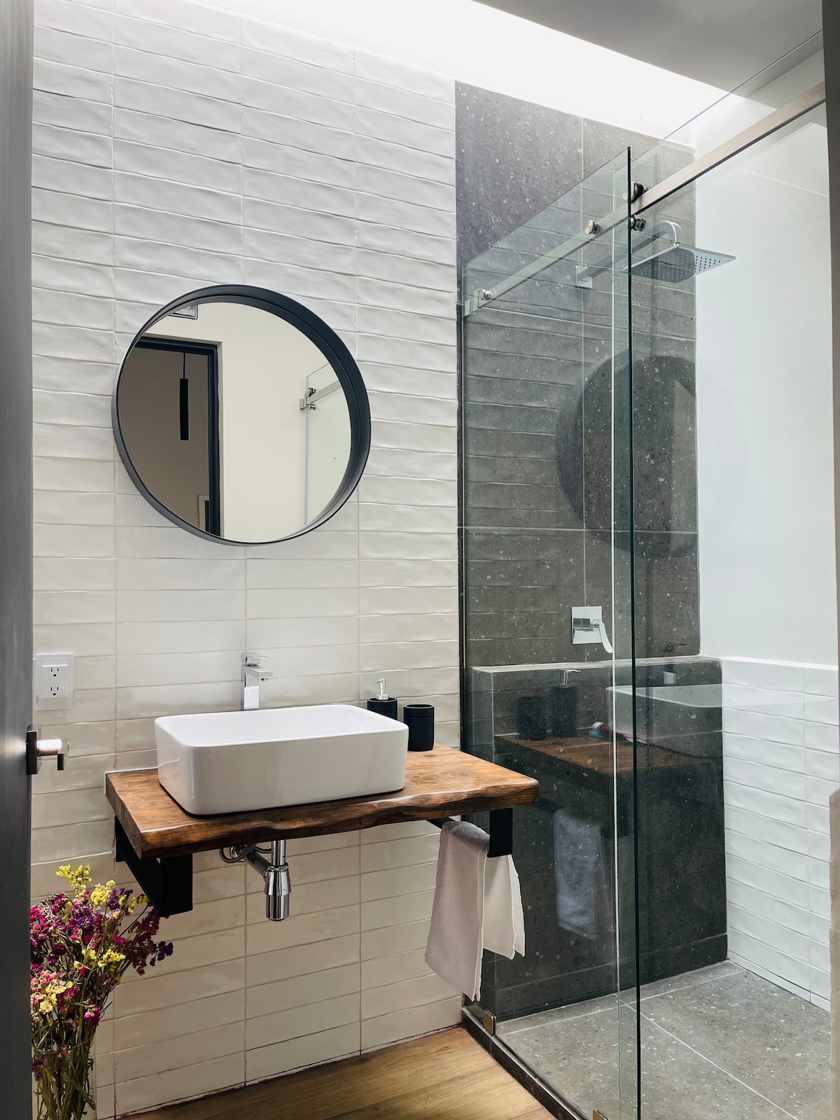 A stylish bathroom features a round mirror above a modern sink placed on a wooden countertop. A glass shower enclosure is visible with sleek tile work, and natural light enhances the clean lines and contemporary design.