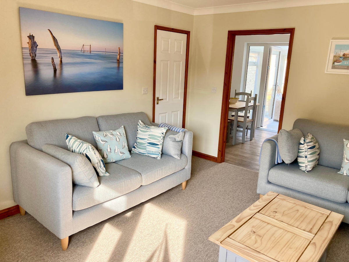 A comfortable living area features two light grey sofas adorned with decorative cushions. A wooden coffee table is positioned centrally, and a large artwork depicting a coastal scene hangs on the wall. Natural light floods in from a doorway leading to the conservatory.