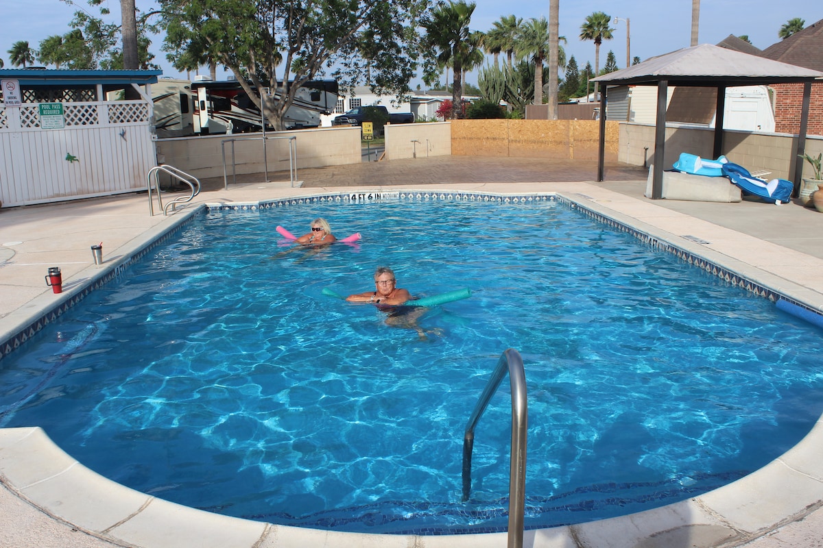 A sparkling swimming pool is bordered by a clean deck, featuring comfortable lounge areas under shaded structures. Two individuals enjoy the clear water with colorful flotation devices. A backdrop of palm trees and well-maintained landscaping enhances the inviting atmosphere.