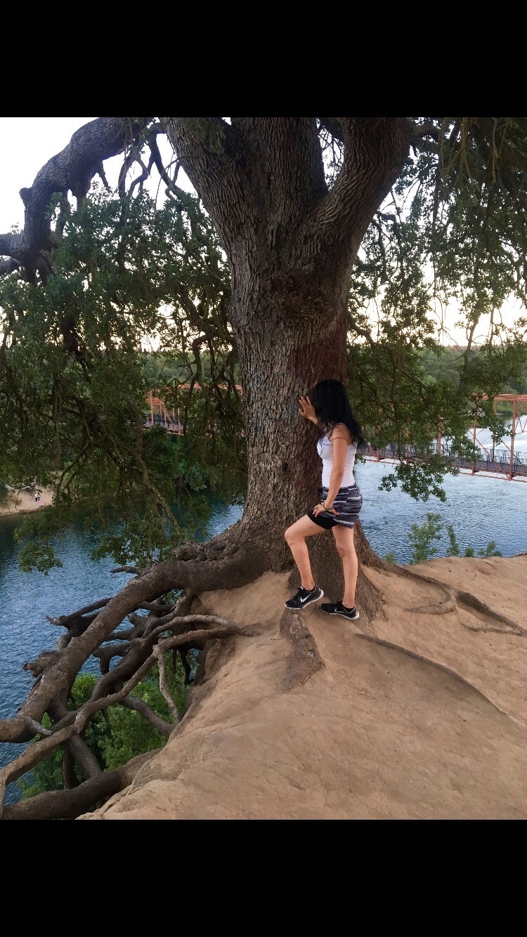 An individual stands on a rocky ledge next to a large tree with sprawling roots, overlooking a serene body of water. The landscape features a gently sloping bank, enhancing the natural setting, while soft light highlights the contours of the terrain.