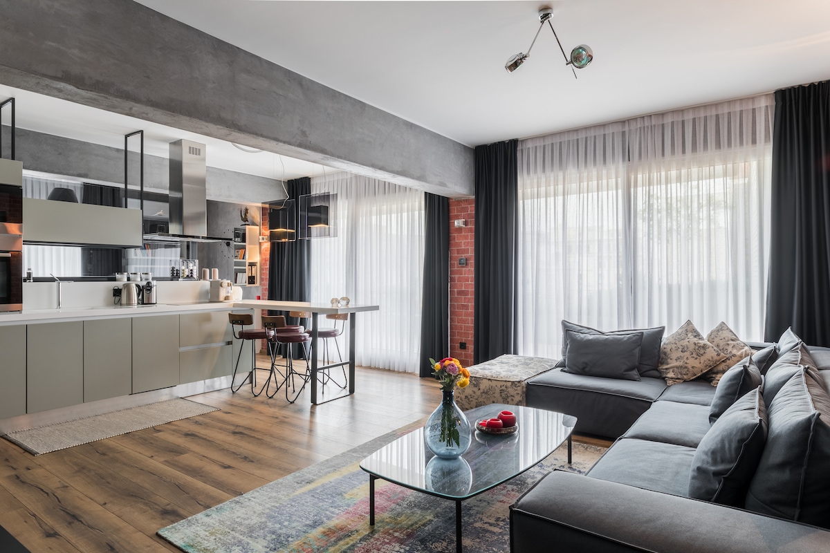 An open living area showcases a modern kitchen with an island bar and seating. Natural light floods through large windows, creating a bright atmosphere. A spacious sectional sofa, adorned with cushions, faces a low glass coffee table, enhancing the laid-back vibe of the space.