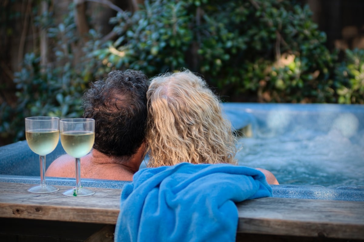 Two guests are seated in a hot tub, surrounded by lush greenery. A blue towel is draped over the edge, and two glasses filled with white wine rest on a ledge nearby, suggesting a relaxed atmosphere. The focus is on the soothing setting and the guests enjoying their time.