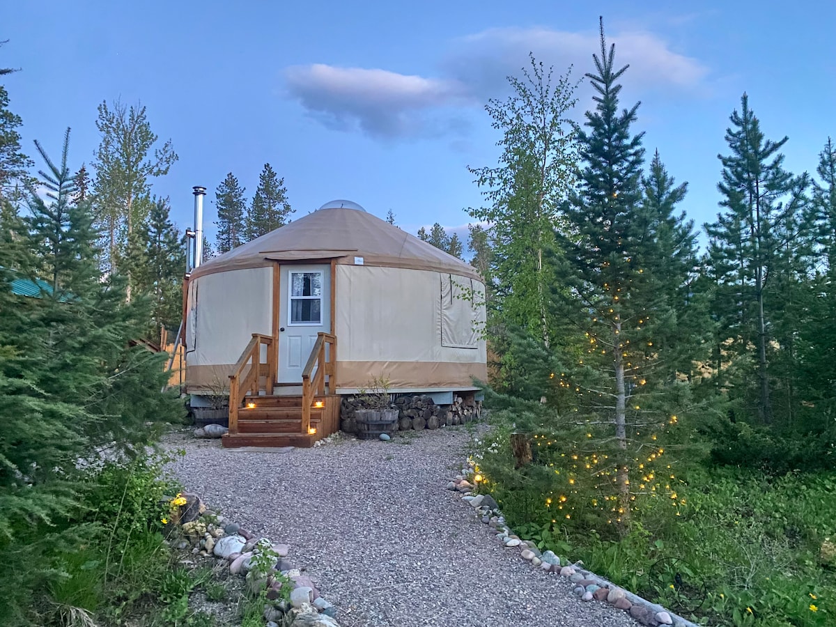 A cozy yurt is nestled among trees, with a welcoming wooden staircase leading to the entrance. Soft lighting lines the path, illuminating the rocky walkway and enhancing the natural surroundings as twilight approaches.