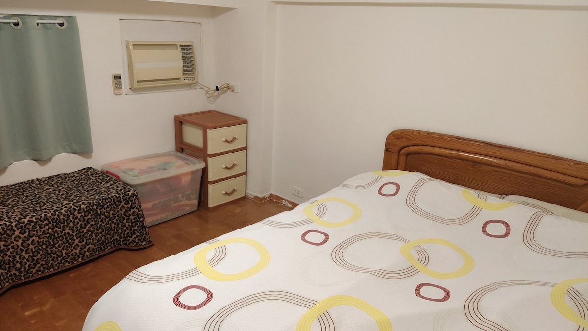 A simple room features a spacious bed with a patterned duvet. A small bedside table stands beside the bed, while a storage box with a cover is located at the foot of the bed. A window air conditioning unit is mounted on the wall.