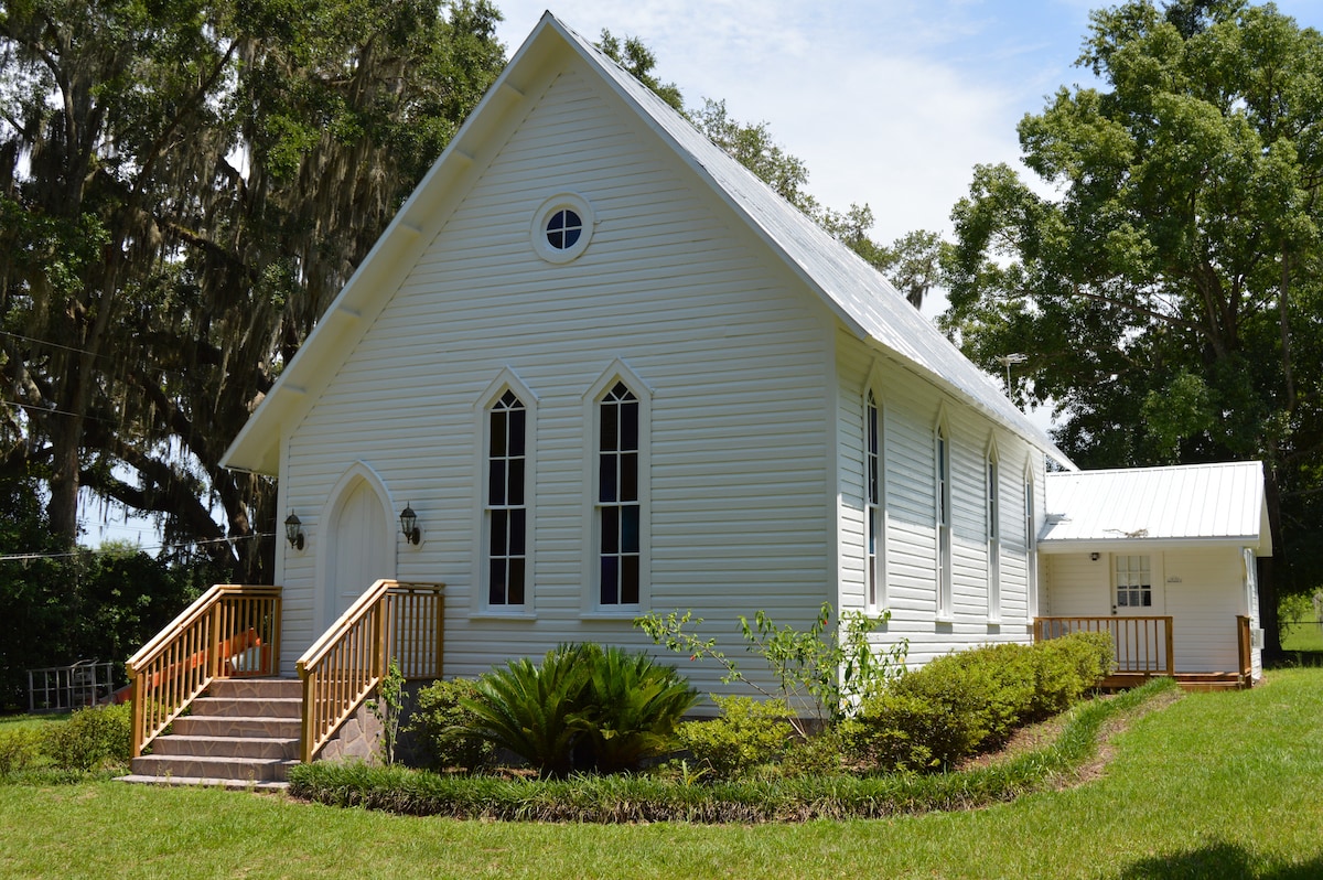 The exterior of a charming white church features Gothic-style windows and a pointed roof, surrounded by lush greenery. A set of wooden steps leads to the main entrance, complemented by well-maintained landscaping, creating a serene setting in the Old South.