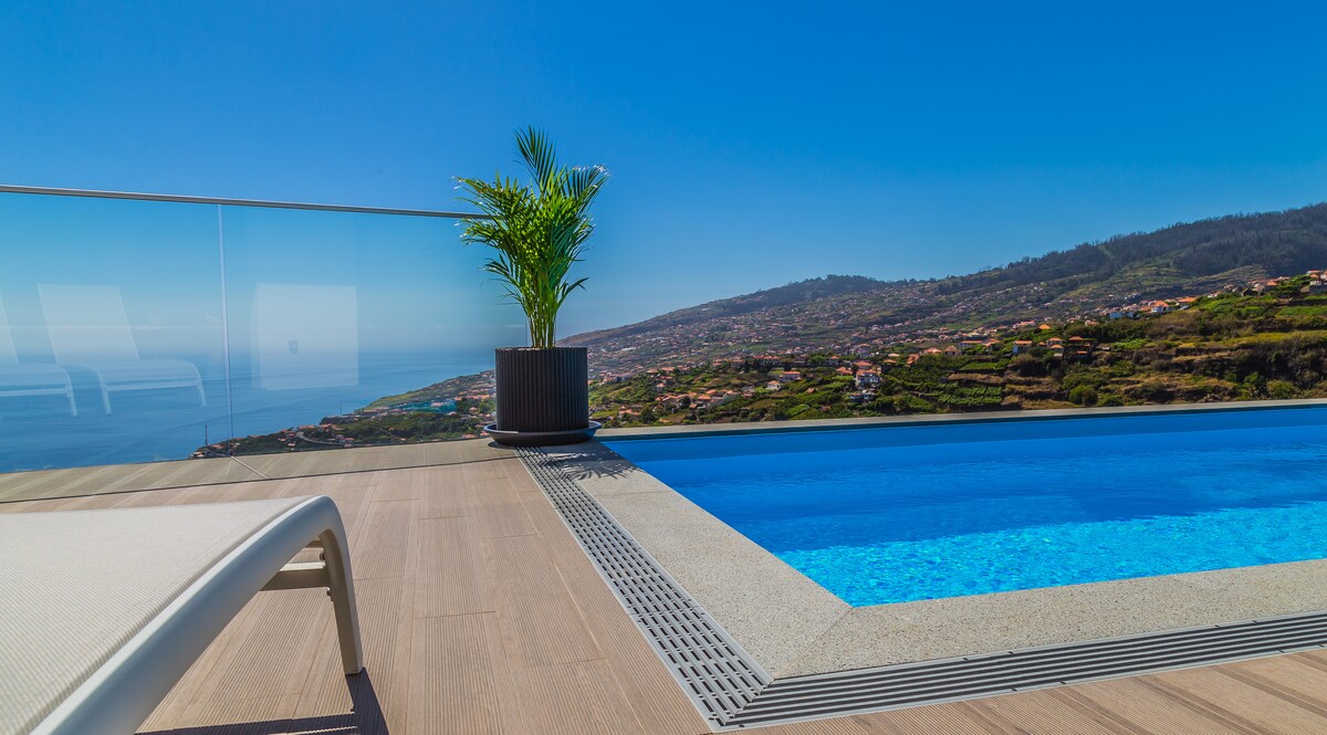 A private heated pool is situated on a sunlit terrace, offering panoramic views of the ocean and surrounding hills. A potted plant adds a touch of greenery beside the pool, while clear blue skies enhance the serene atmosphere.