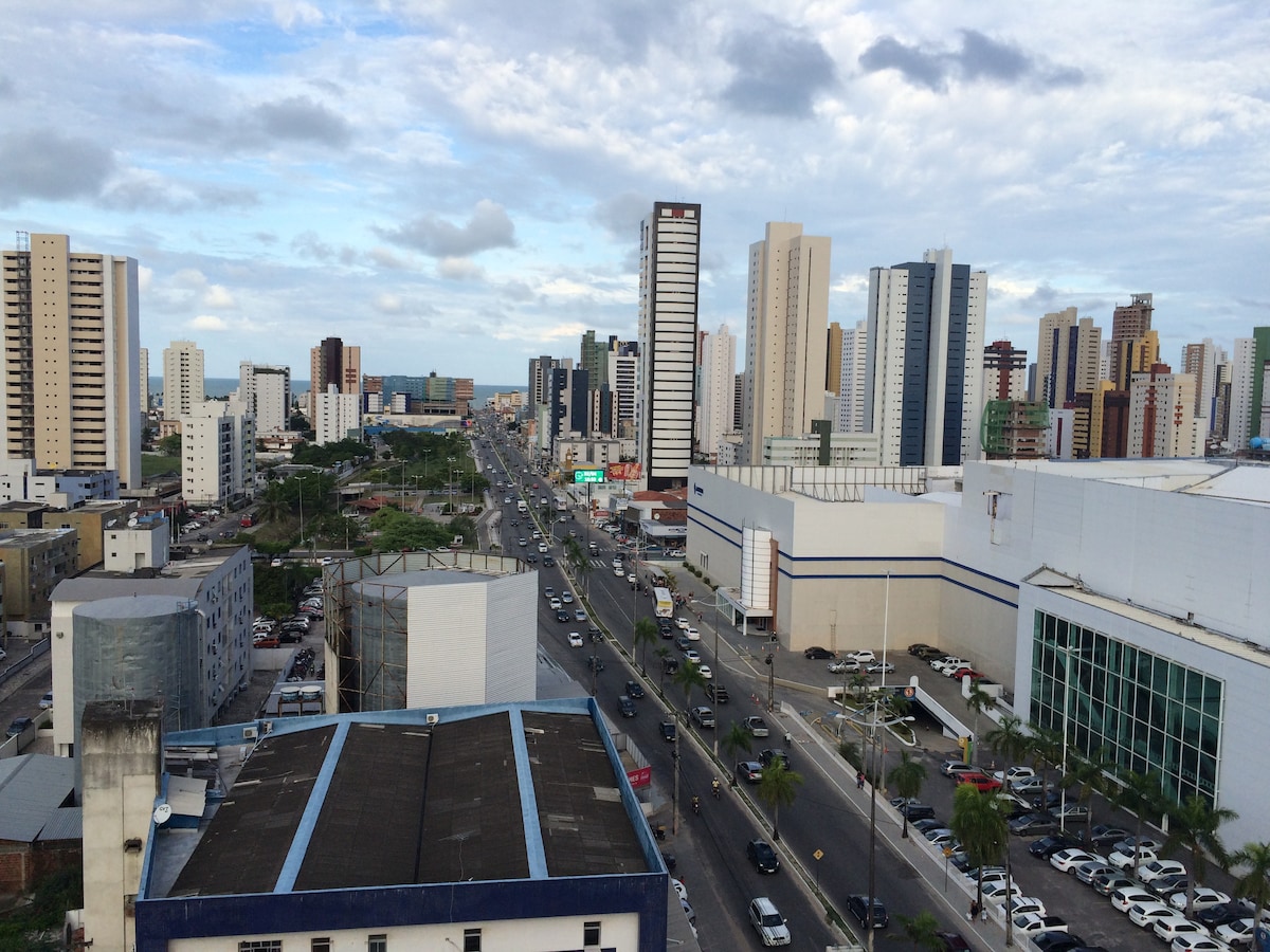A panoramic view of a bustling urban landscape is presented, showcasing numerous high-rise buildings along a main avenue. The scene includes a mix of residential and commercial structures, with trees lined along the roads, and vehicles in motion, indicative of city life.
