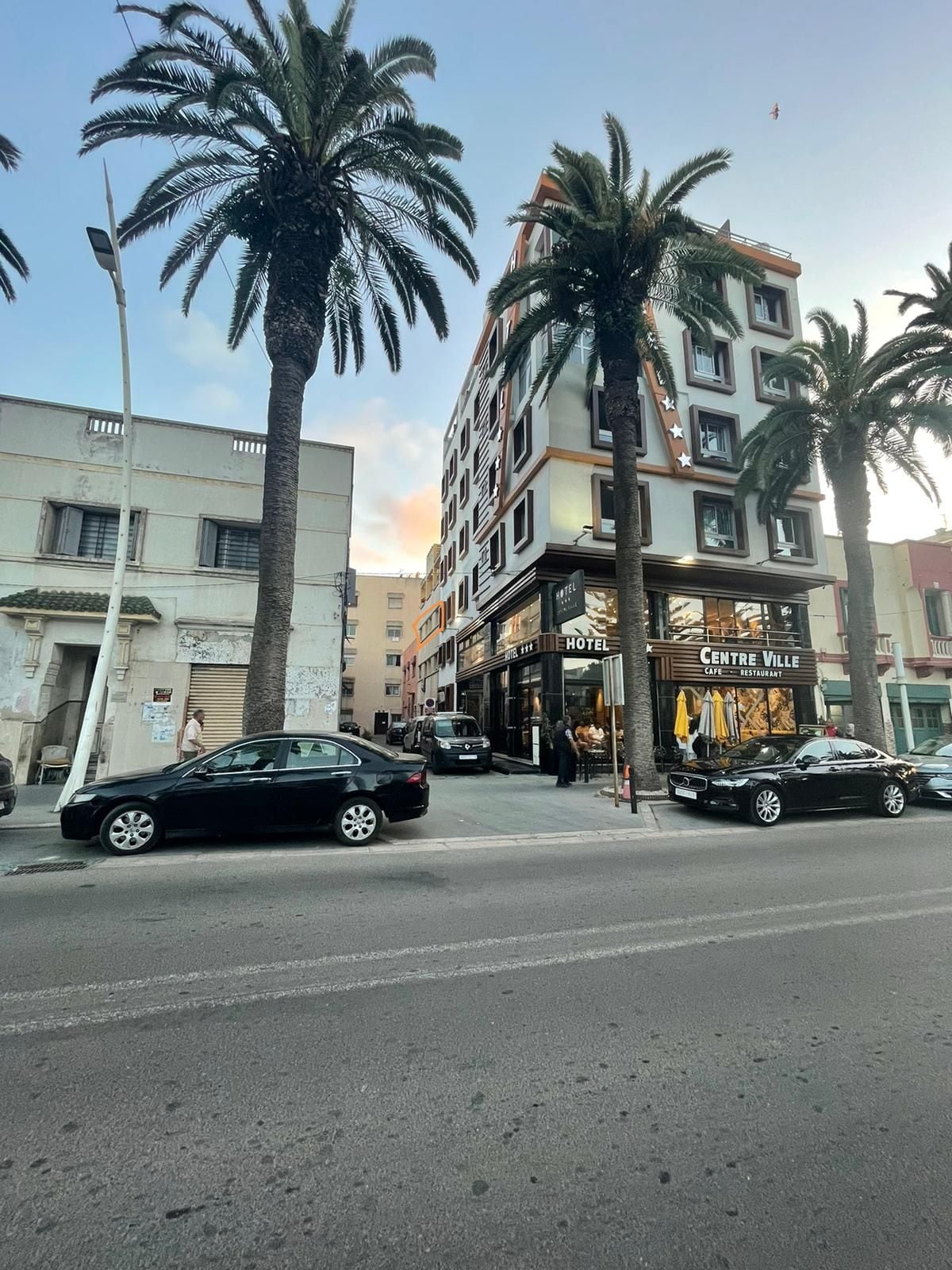 A modern hotel building is seen from the street, surrounded by tall palm trees. The ground level features expansive glass windows displaying the entrance, while parked cars line the street in front. The multi-story structure exhibits a contemporary architectural design.