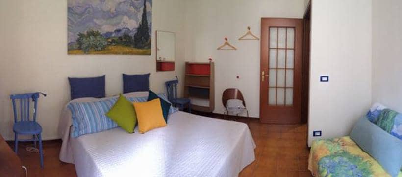 A spacious room features a double bed covered with a light quilt, complemented by colorful pillows. Two blue chairs are positioned near a small table, while a couch with a floral pattern is present. The decor includes a vibrant wall painting and a wooden closet with hangers.