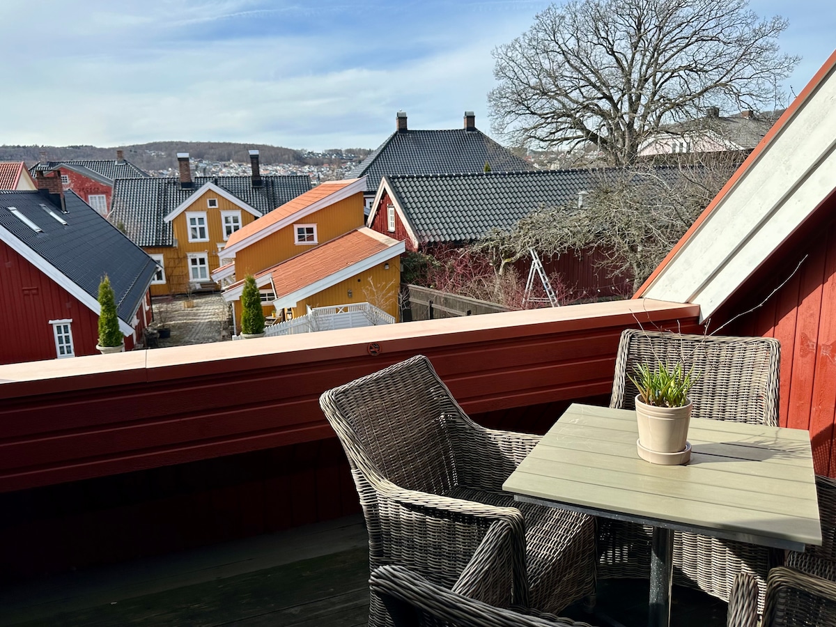 A spacious terrace is shown, featuring a small table and two woven chairs. A potted plant rests on the table. In the background, a view of colorful houses and rooftops is visible, with trees and hills creating a serene backdrop.