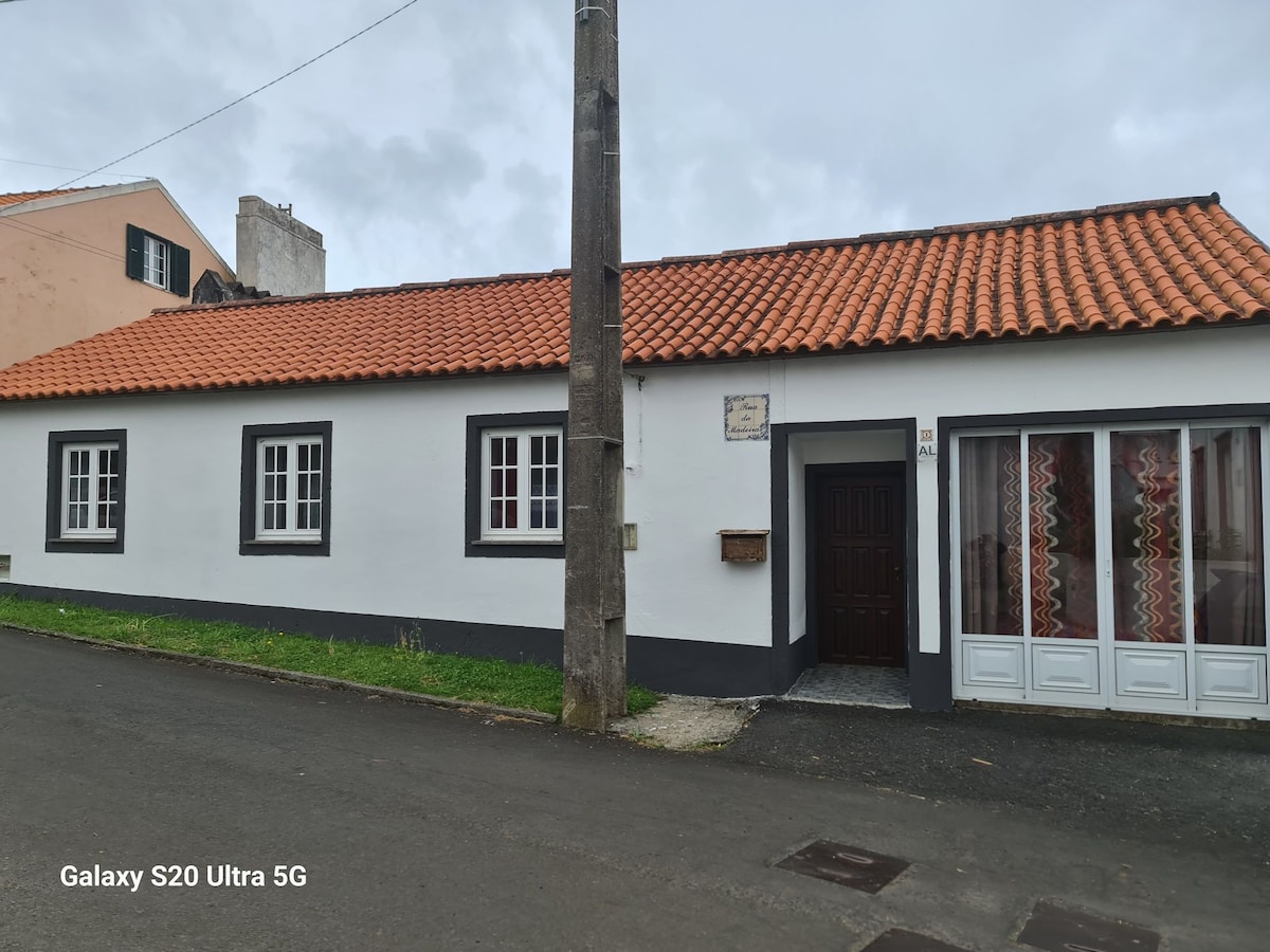 A single-story house with a red-tiled roof is depicted, featuring white walls and dark window frames. The front entrance is characterized by a decorative stone plaque and a glass door adjacent to patterned curtains. The surrounding area includes a quiet street lined with greenery.