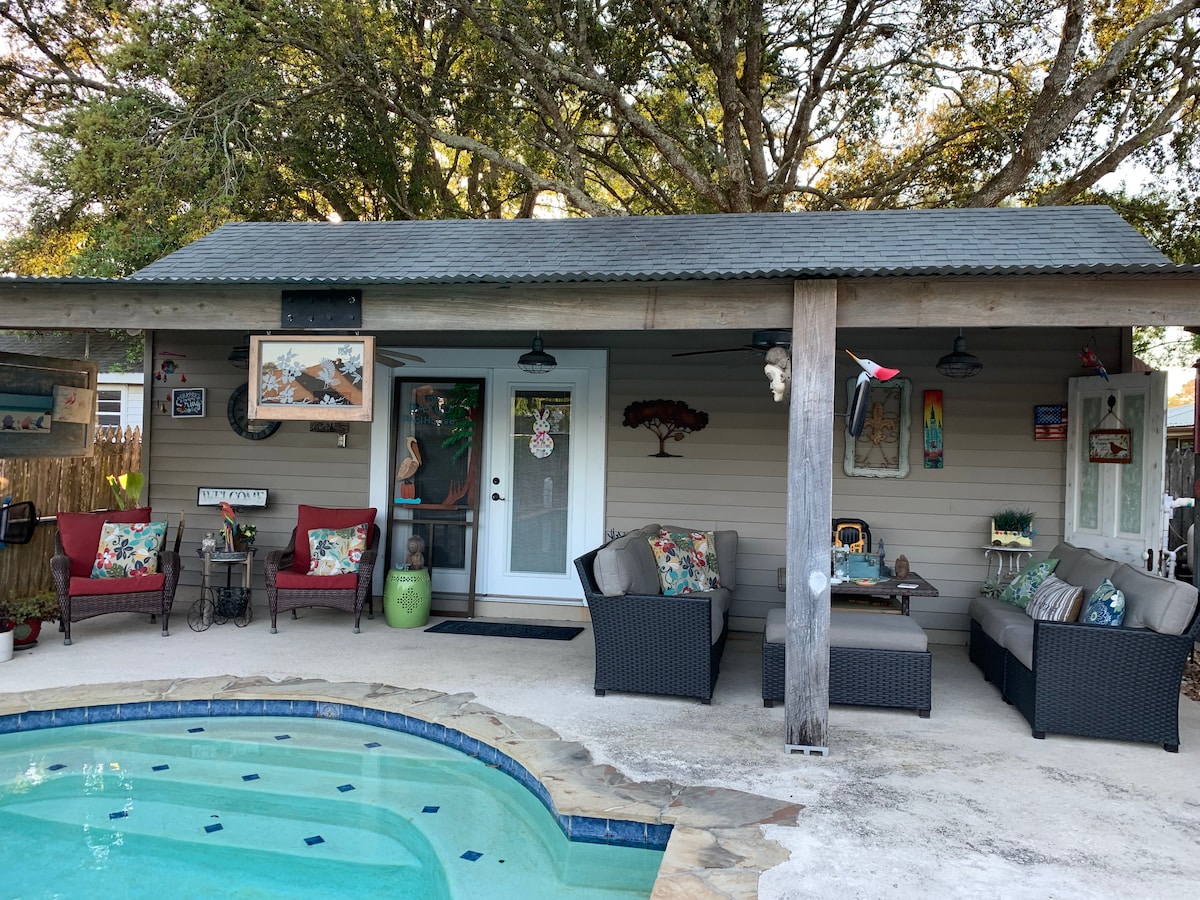 A shaded patio area features multiple seating arrangements, including cushioned couches and vibrant chairs. Decorative art and plants adorn the walls, while a private pool with a stone border complements the space, creating an inviting atmosphere for relaxation.