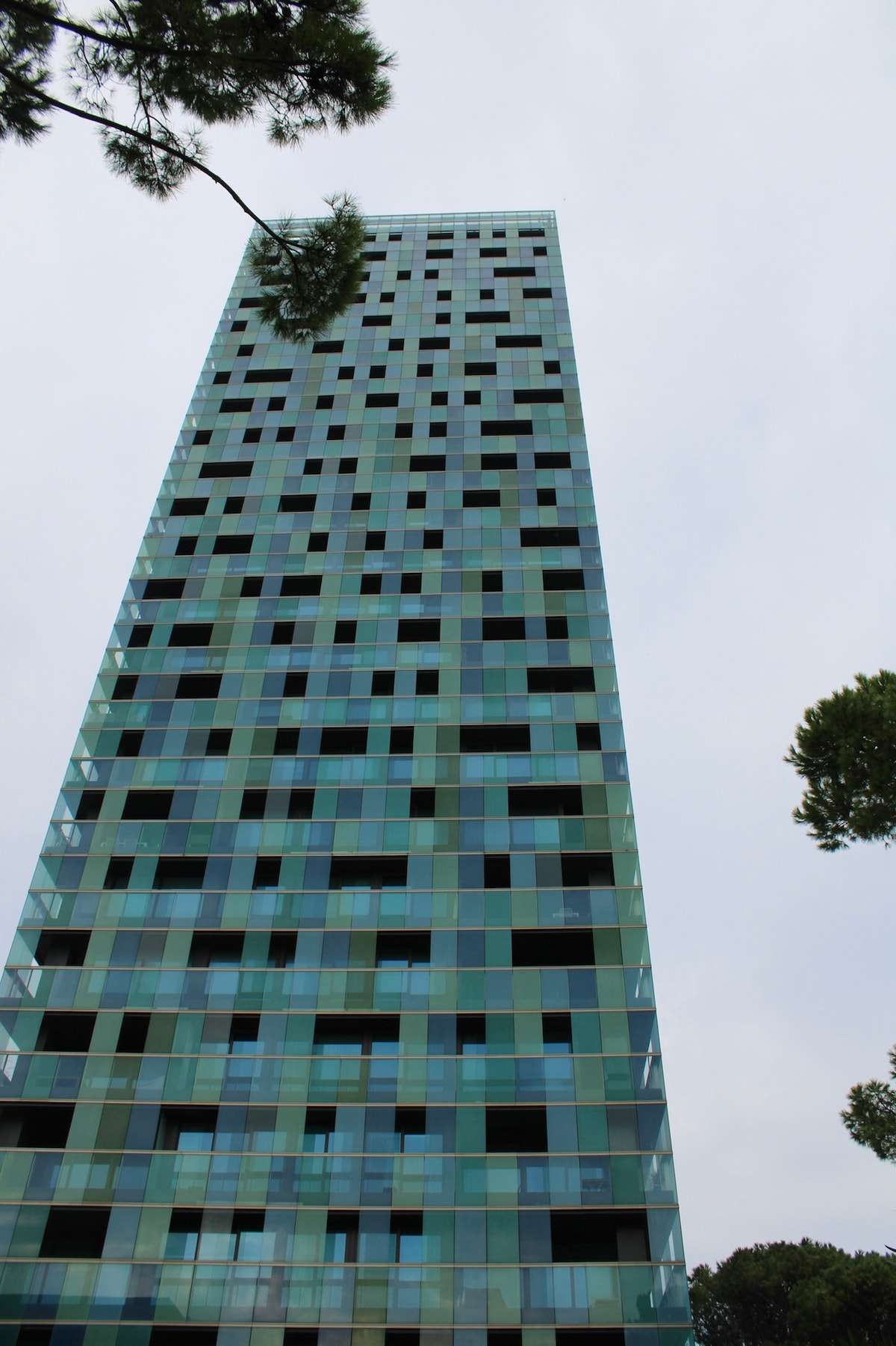 A modern high-rise building with a façade of green and blue glass panels is captured from below. The structure features a grid-like pattern of windows and is surrounded by trees, creating a blend of nature and contemporary architecture.