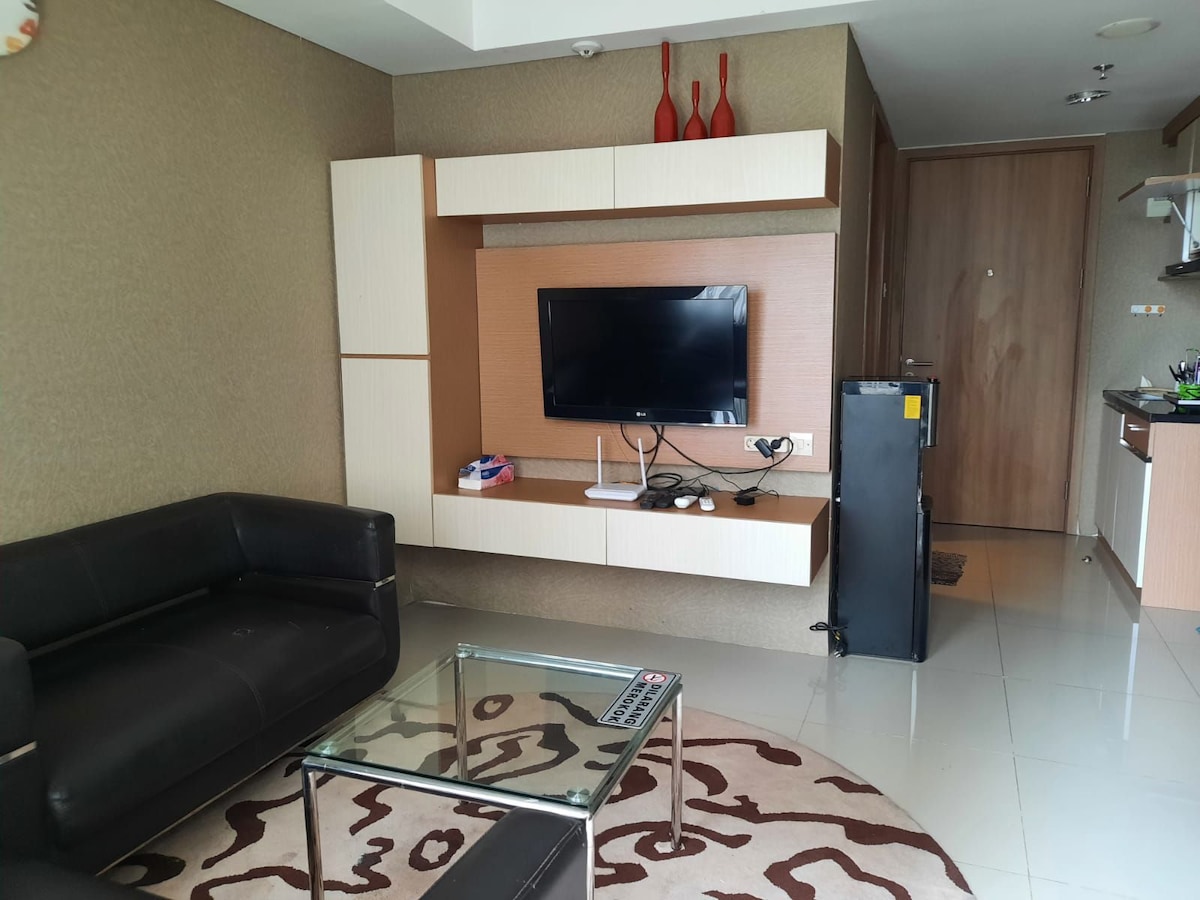 A modern living space features a black sofa and a glass coffee table placed on a patterned rug. A wall-mounted television is positioned above a sleek storage unit. Light-colored cabinetry is seen in the background, leading to the kitchen area, with a door visible to the side.