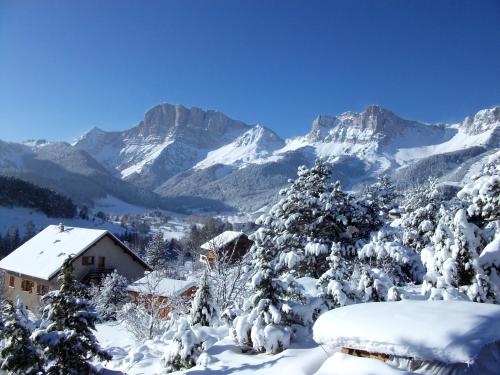 A winter landscape reveals snow-covered mountains and trees under a clear blue sky. The scene includes distant peaks and a few homes nestled among the snow, creating a serene atmosphere. The white snow blankets the ground and rooftops, enhancing the tranquil setting.