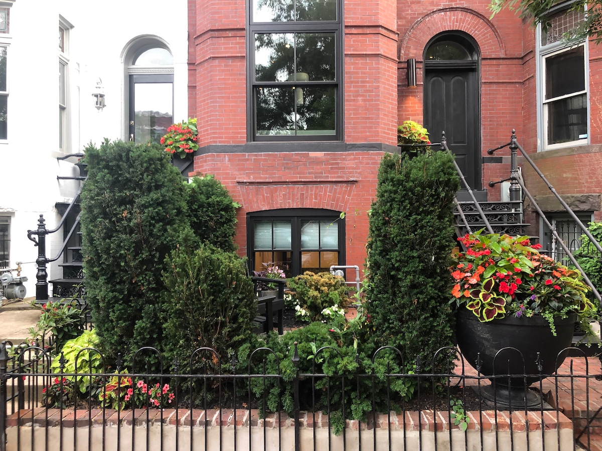 A landscaped front garden features lush greenery and vibrant flowers, bordered by a black wrought iron fence. A small seating area with a table and chairs is visible through the large windows. The inviting brick facade of the building provides a warm backdrop.