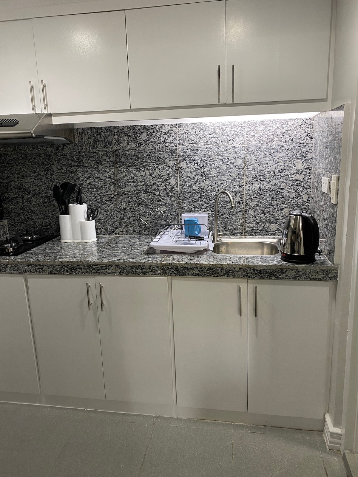 A modern kitchen area is presented with sleek white cabinetry and a granite countertop. Essential appliances such as a kettle and sink are visible, along with neatly arranged utensils. The space is well-lit, enhancing the functionality of the cooking area.