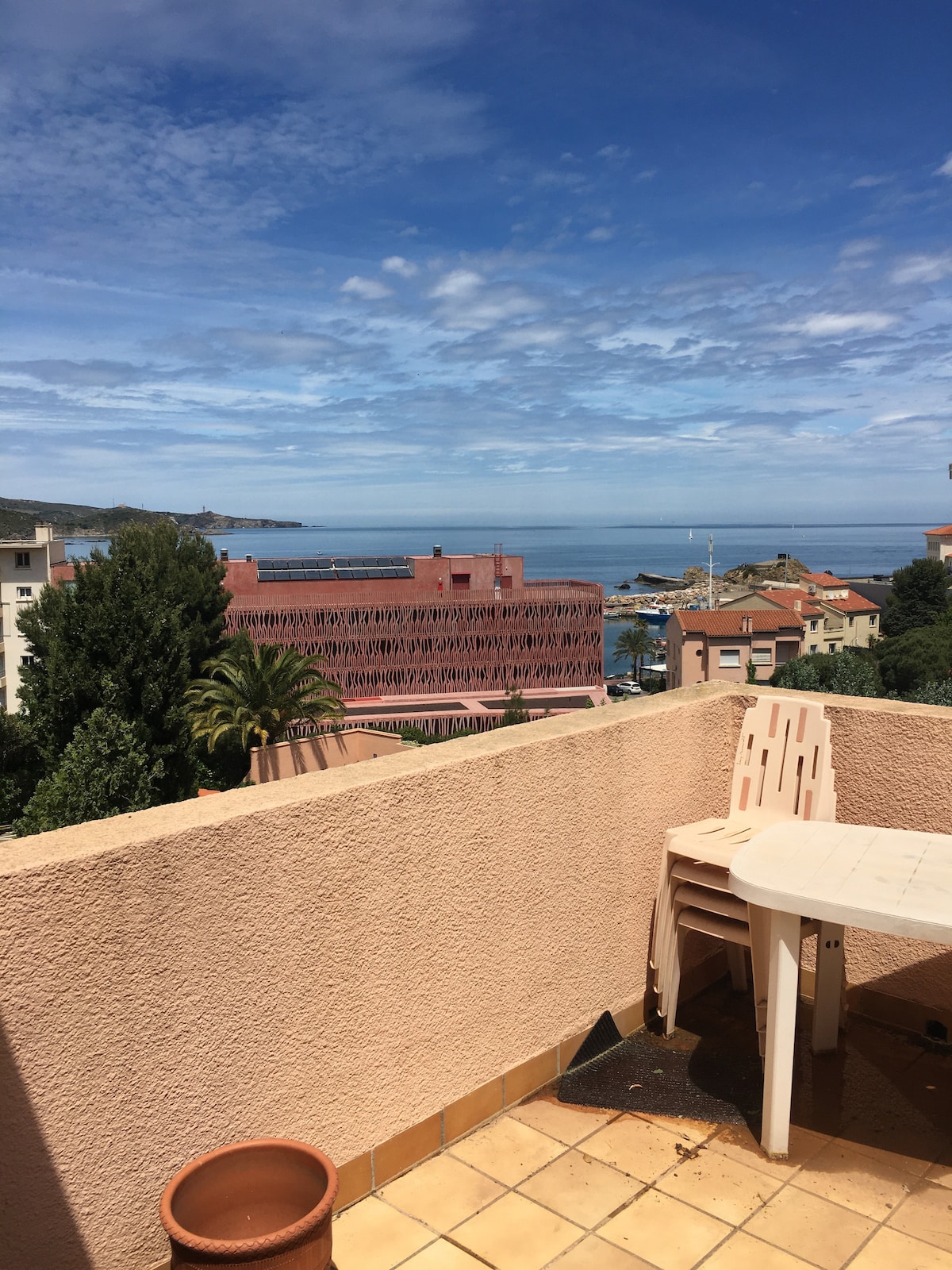 A terrace is seen with a table and stacked chairs, framed by a low wall. The ocean is visible in the distance, accompanied by a clear sky and scattered clouds. Buildings and palm trees are present in the foreground, contributing to a relaxed coastal setting.