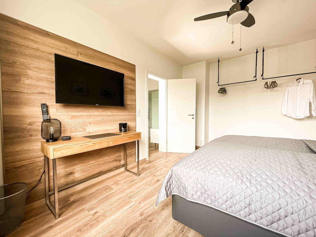 A spacious bedroom is furnished with a plush bed and a stylish wooden accent wall. A wall-mounted flat-screen TV and a small desk with a coffee maker are included. Industrial-style clothing racks are featured, along with a ceiling fan to enhance airflow.